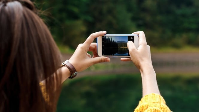 Seorang wanita sedang  memotret pemandangan melalui smartphone