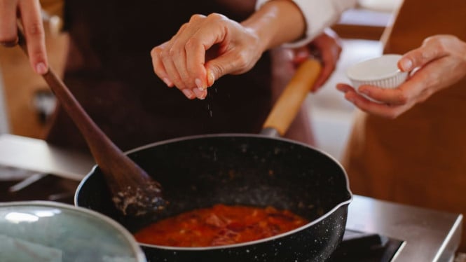 2 orang yang sedang memasak dan salah satunya menambah bumbu masak